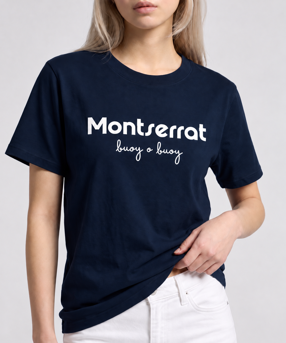 A woman wearing a navy blue Montserrat crewneck t-shirt with 'Montserrat buoy o buoy' printed in white across the chest, paired with white pants, against a white background.