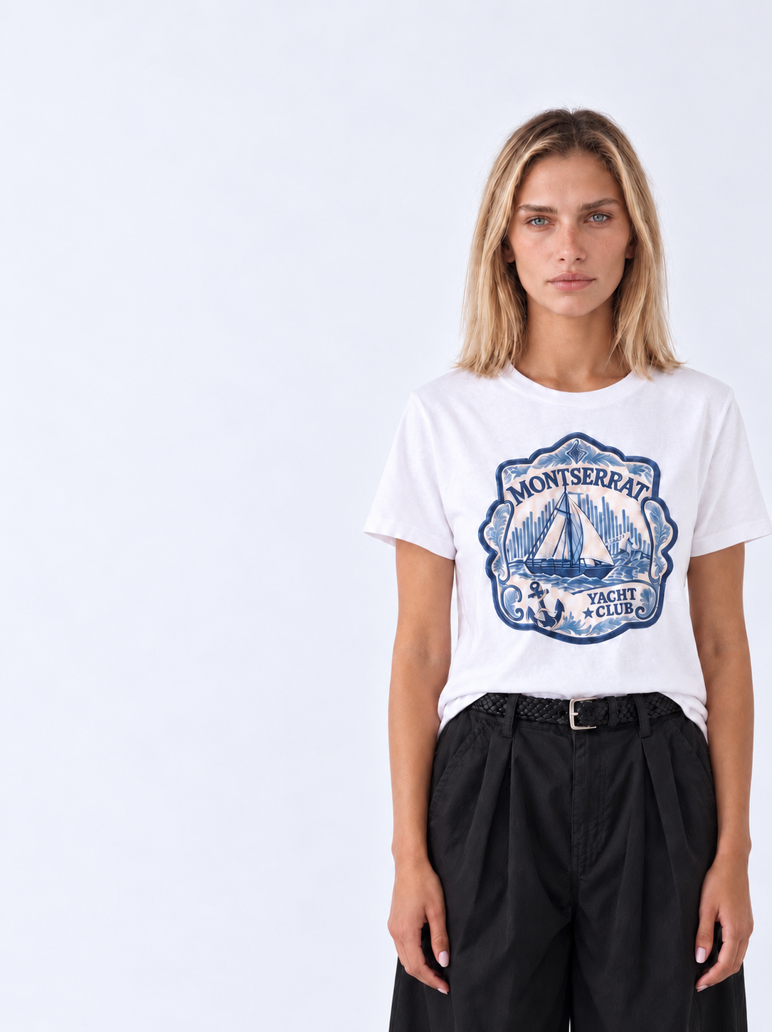 A woman modeling a white short-sleeve t-shirt with a blue vintage-style 'Montserrat Yacht Club' graphic on the front, paired with black trousers, against a plain light gray background.