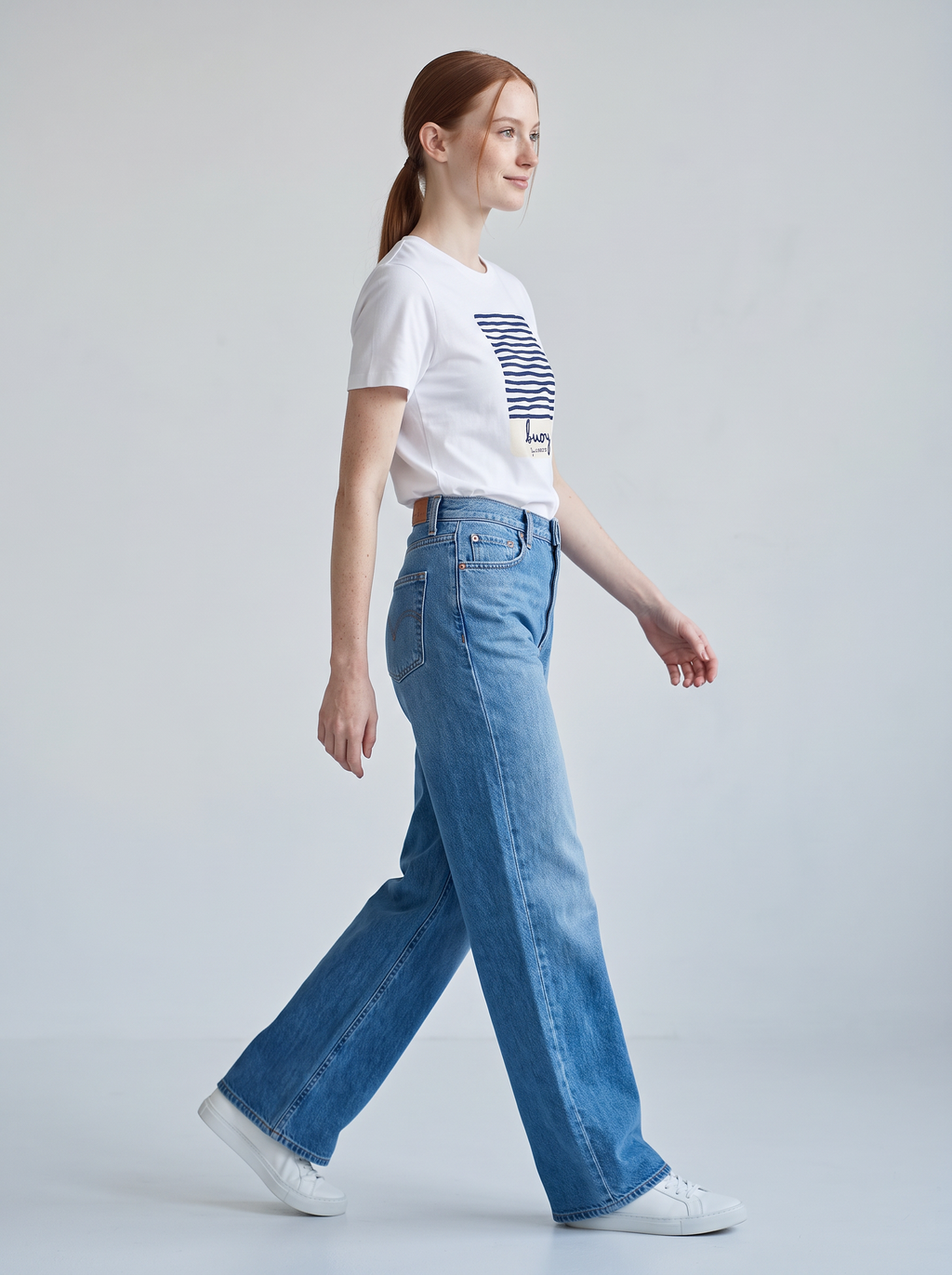 A woman in a white graphic Montserrat t-shirt and blue wide-leg jeans walking in profile against a light grey studio background.